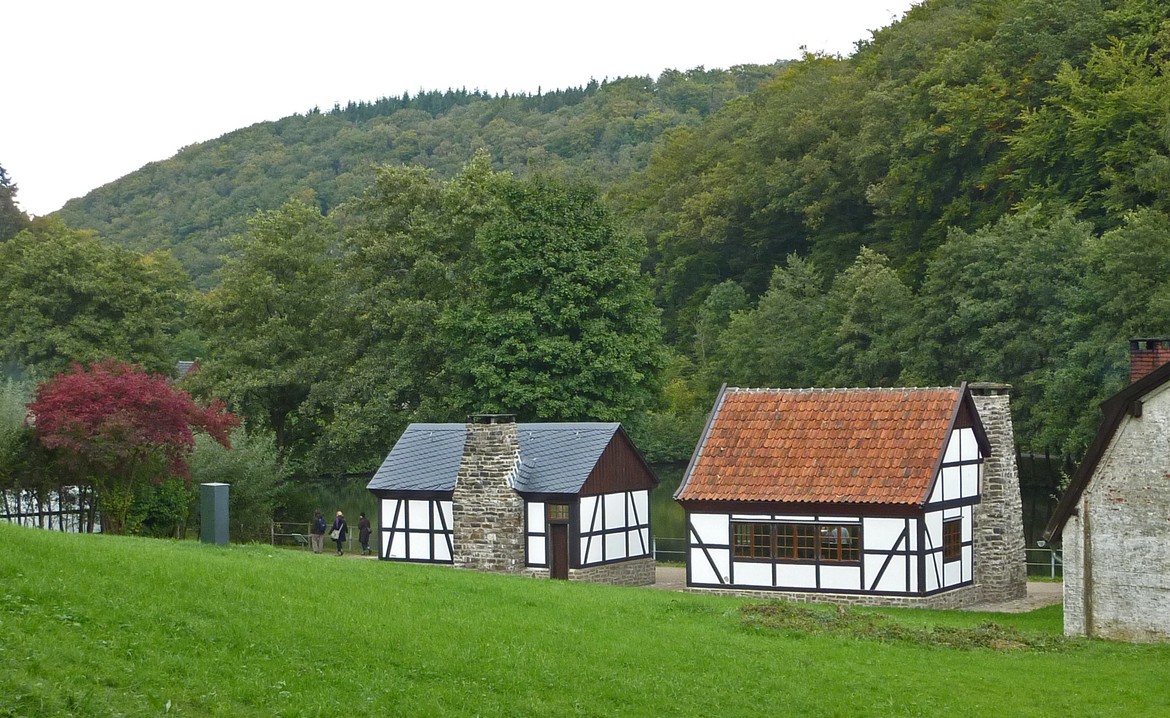 Im Vordergrund ist eine Wiese auf der 2 kleine Fachwerkhäuser stehen. Im Hintergrund ist dichter Laubwald.